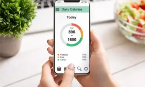Dłoń trzyma smartfon z aplikacją "Daily Calories", pokazującą spożycie 896 z 1600 kcal. Widoczny jest wzór mifflina w rozkładzie białek, tłuszczów i węglowodanów.