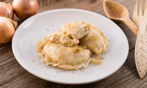 Pyszne pierogi z kapustą i grzybami, posypane pieprzem, na białym talerzu. Obok cebula i drewniane sztućce.