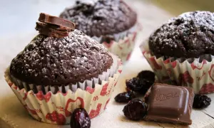 Czekoladowe muffiny posypane cukrem pudrem, ozdobione kawałkami czekolady E. Wedel. Obok leżą suszone wiśnie. Pyszne, choć kaloryczne!