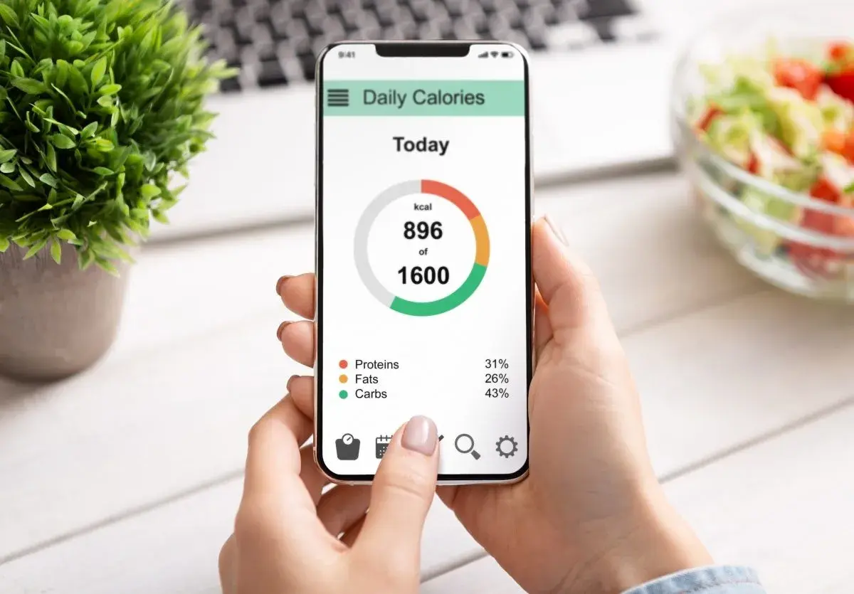 Dłoń trzyma smartfon z aplikacją "Daily Calories", pokazującą spożycie 896 z 1600 kcal. Widoczny jest wzór mifflina w rozkładzie białek, tłuszczów i węglowodanów.