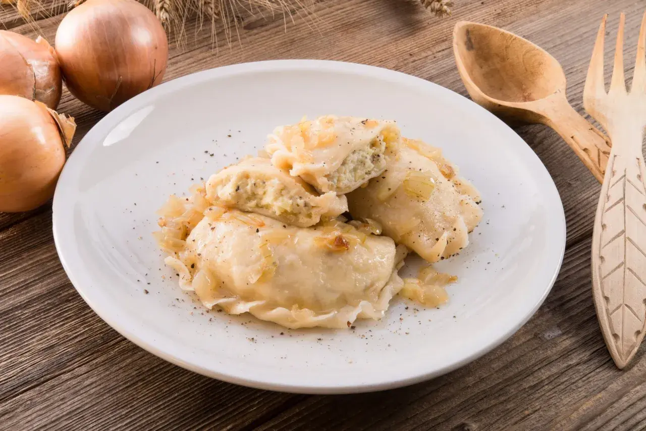 Pyszne pierogi z kapustą i grzybami, posypane pieprzem, na białym talerzu. Obok cebula i drewniane sztućce.