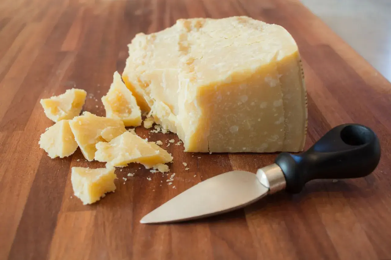 Kawałek parmezanu i kilka mniejszych kawałków na desce. Obok leży nóż do sera. Warto sprawdzić parmezan kcal przed spożyciem.