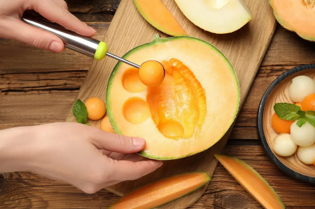 Ręka formuje kulki z melona, który ma niski melon indeks glikemiczny. Obok leżą kawałki melona i miseczka z owocowymi kulkami.